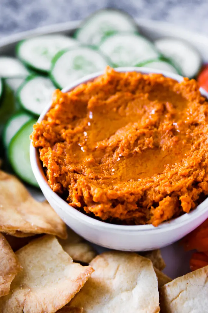 a close up of a bowl of roasted carrot hummus next to pita chips and sliced cucumbers