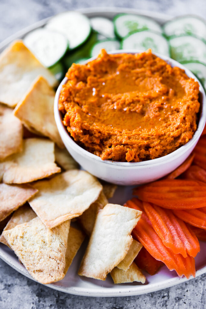 a plate of pita chips, carrot chips, and cucumber slices with a bowl of roasted carrot hummus