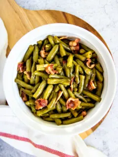 a round, white baking dish with green bean and bacon casserole on a round wooden cutting board