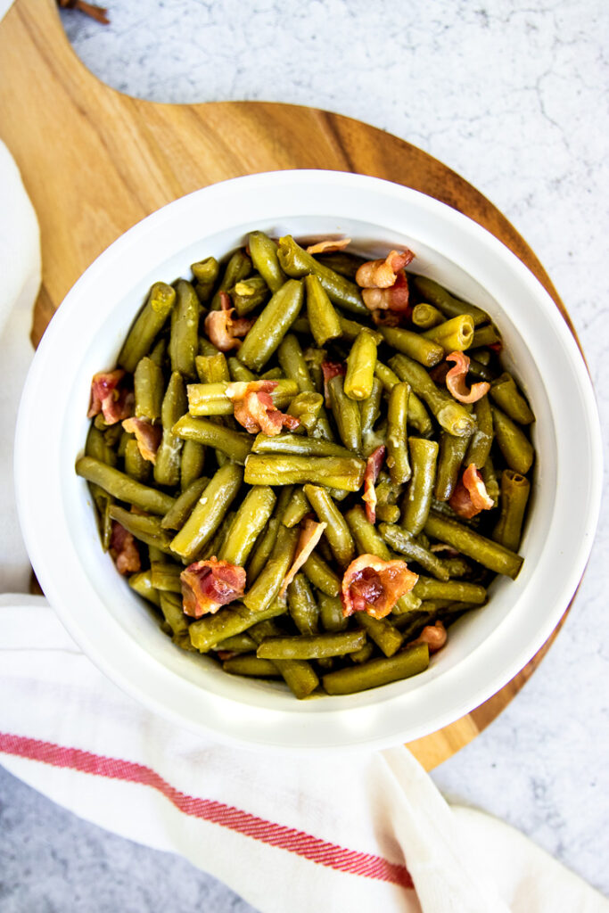 a round, white baking dish with green bean and bacon casserole on a round wooden cutting board