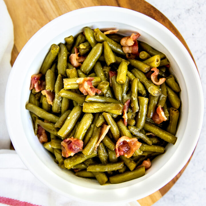 a round, white baking dish with green bean and bacon casserole on a round wooden cutting board