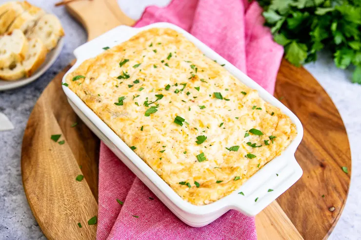 a lower angle of the crab dip in a white rectangular baking dish on a circular wooden board on a red napkin with parsley in the background