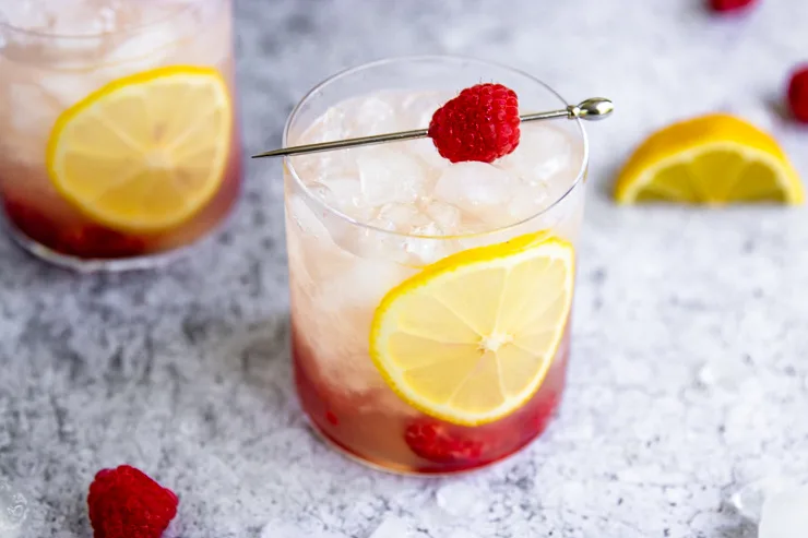 a rocks glass filled with crushed ice and a slice of lemon topped with a metal pick with a raspberry