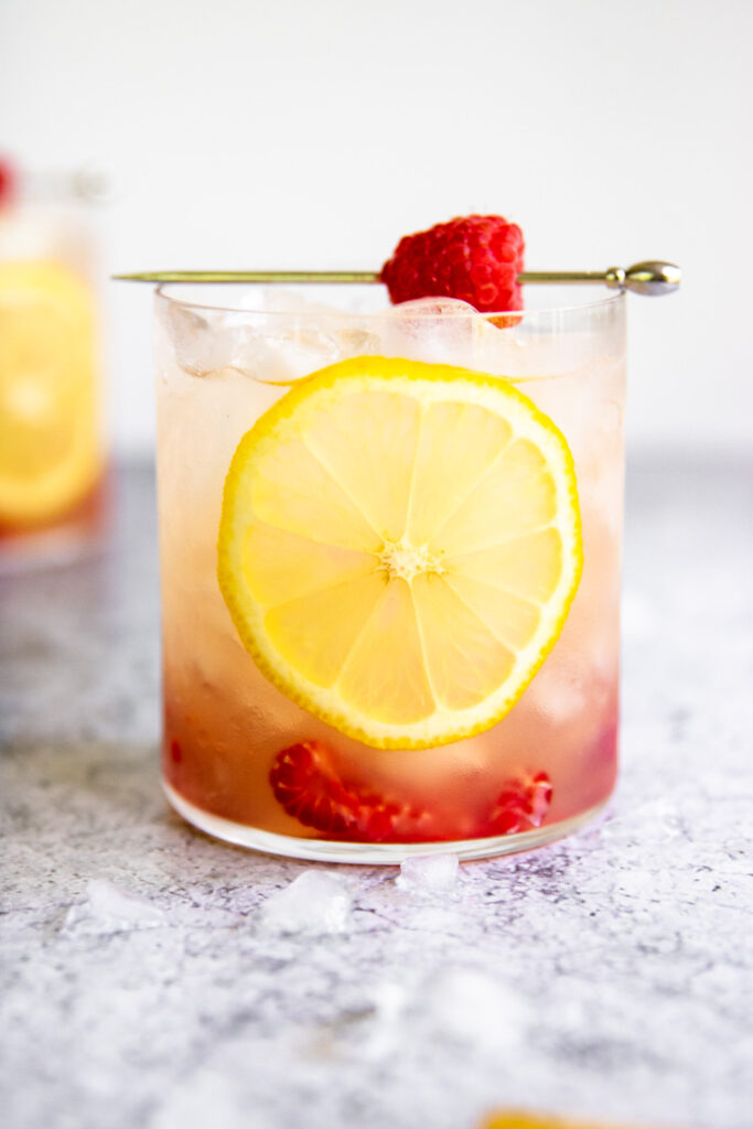 a glass of raspberry bramble with a lemon round against the glass and a metal pick with a raspberry sitting on top