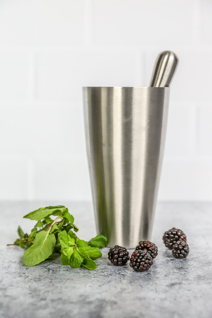 a metal shaker and muddler, mint leaves, and fresh blackberries sitting next to each other