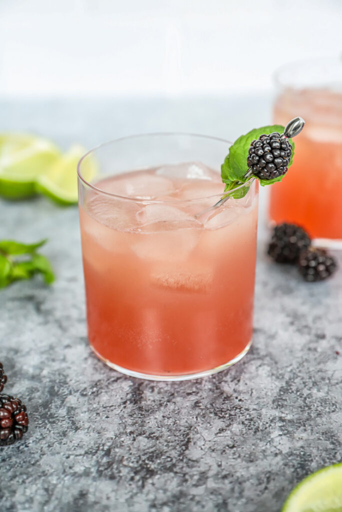 a rocks glass with light pink blackberry bourbon smash with a blackberry and mint leaves on a metal cocktail pick