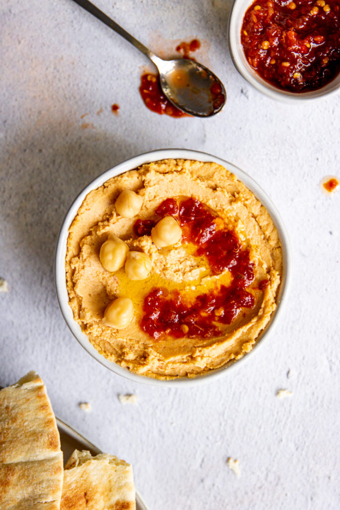 a plate of pita, spoon, and bowl of sambal oelek surrounding the bowl of hummus