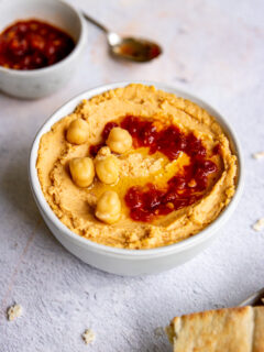 a white bowl of hummus with chick peas and sambal oelek on top and a small bowl of red sambal oelek in the back