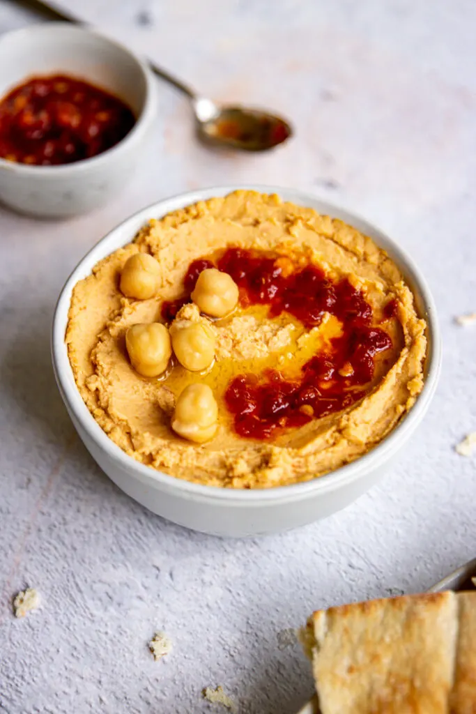 a white bowl of hummus with chick peas and sambal oelek on top and a small bowl of red sambal oelek in the back