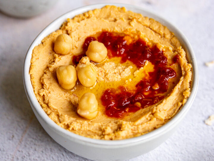 a white bowl of hummus with chick peas and sambal oelek on top and a small bowl of red sambal oelek in the back