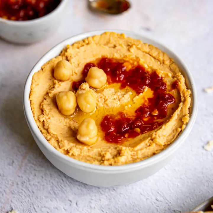 a white bowl of hummus with chick peas and sambal oelek on top and a small bowl of red sambal oelek in the back