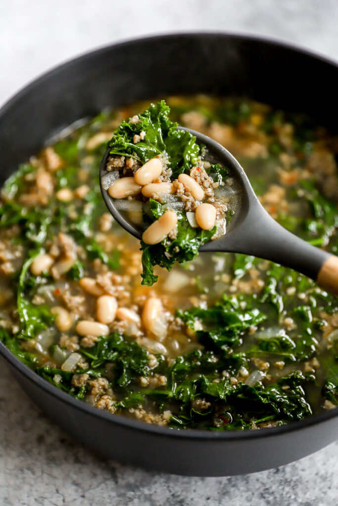 a grey ladle scooping out sausage kale white bean soup over the pot