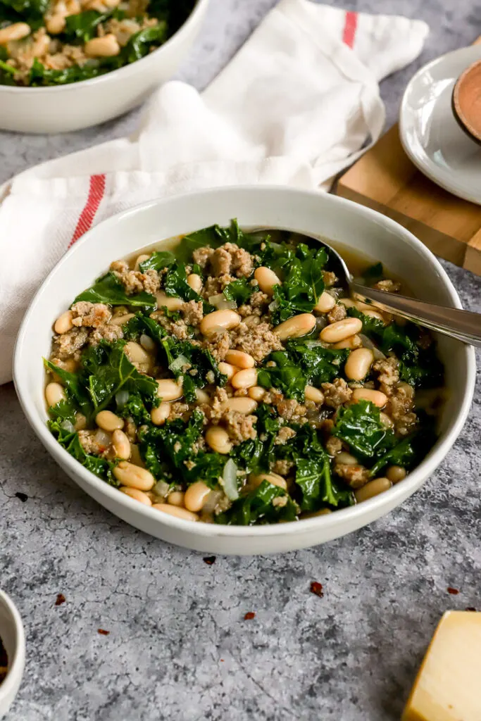 a close up of a white bowl of sausage kale white bean soup with a spoon