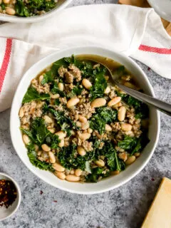 a white bowl of sausage kale white bean soup next to a white napkin with a red stripe