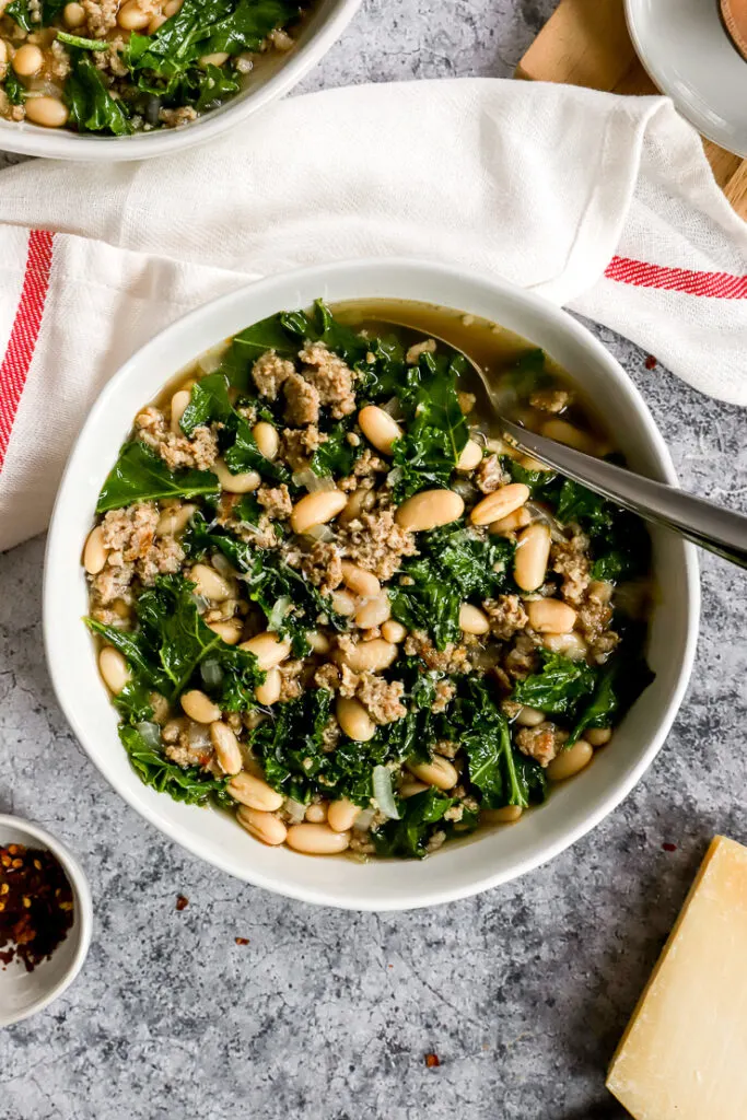 a white bowl of sausage kale white bean soup next to a white napkin with a red stripe