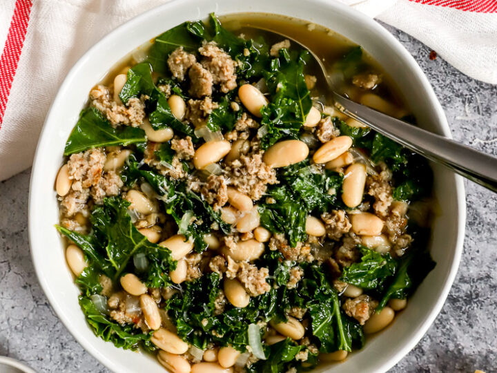 a white bowl of sausage kale white bean soup next to a white napkin with a red stripe