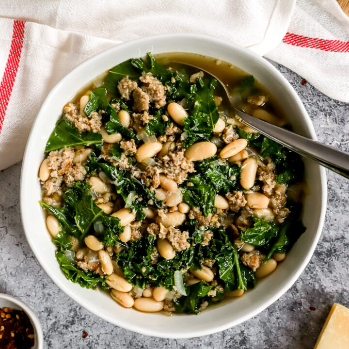 a white bowl of sausage kale white bean soup next to a white napkin with a red stripe