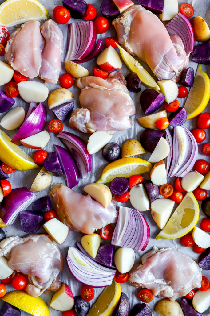 a close up of raw chicken thighs, red onion slices, lemon slices, tomatoes, and cut potatoes