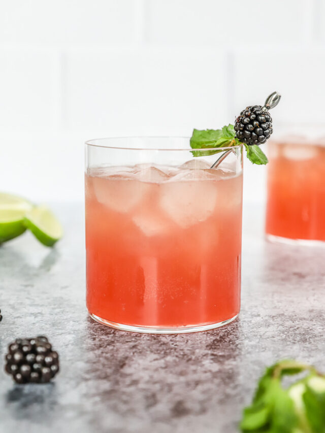 a blackberry bourbon smash in a rocks glass with mint leaves and blackberry on a metal cocktail pick and mint and blackberries scattered