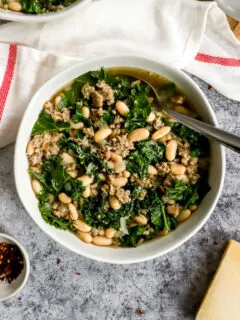 a white bowl of sausage kale white bean soup next to a white napkin with a red stripe