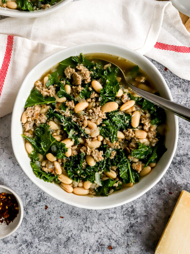 a white bowl of sausage kale white bean soup next to a white napkin with a red stripe