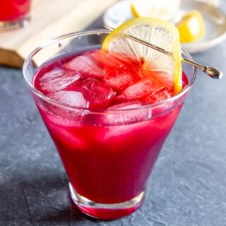 a cosmopolitan glass with cranberry bourbon cocktail and a lemon slice on a cocktail pick on a dark grey background