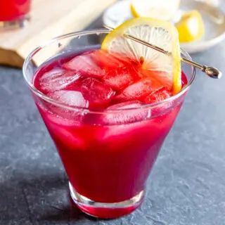a cosmopolitan glass with cranberry bourbon cocktail and a lemon slice on a cocktail pick on a dark grey background