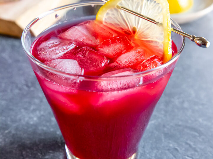 a cosmopolitan glass with cranberry bourbon cocktail and a lemon slice on a cocktail pick on a dark grey background