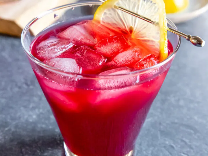 a cosmopolitan glass with cranberry bourbon cocktail and a lemon slice on a cocktail pick on a dark grey background