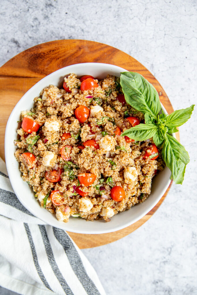 a bowl of italian quinoa salad on a wood cutting board with a napkin and basil garnish