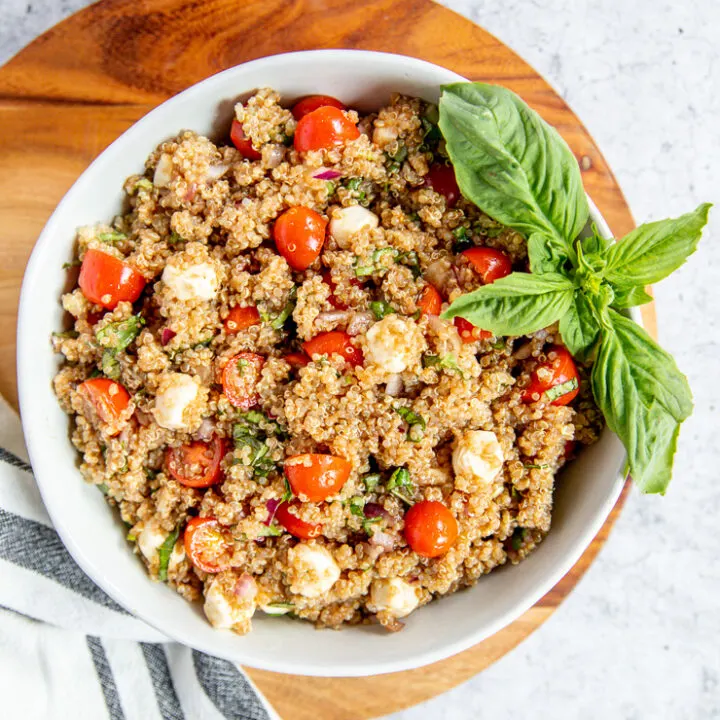 a bowl of italian quinoa salad on a wood cutting board with a napkin and basil garnish