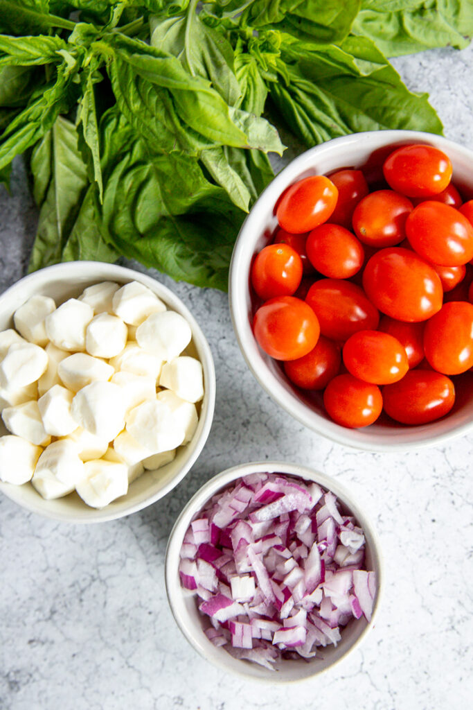 a bunch of basil and three bowls filled with grape tomatoes, pearl mozzarella, and diced red onion