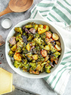 an overhead shot of a bowl of roasted potatoes and broccoli next to a white and green napkin and parmesan grater