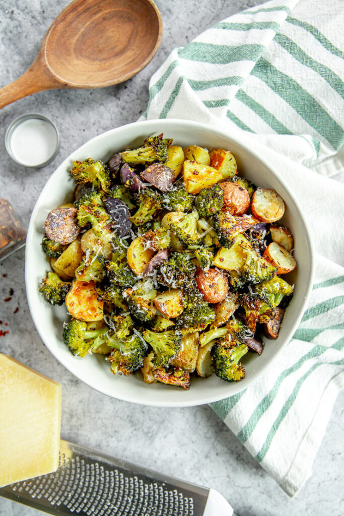 an overhead shot of a bowl of roasted potatoes and broccoli next to a white and green napkin and parmesan grater