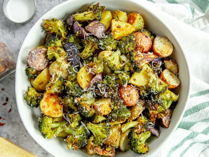 an overhead shot of a bowl of roasted potatoes and broccoli next to a white and green napkin and parmesan grater