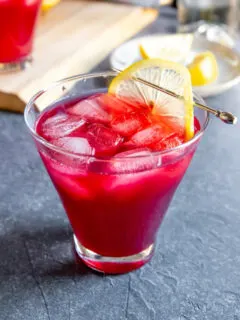 a cosmopolitan glass with cranberry bourbon cocktail and a lemon slice on a cocktail pick on a dark grey background