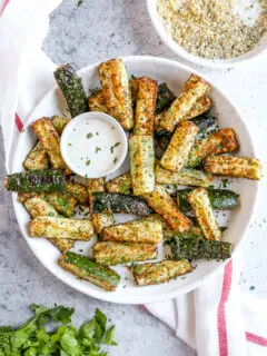 air fried zucchini fries in a large white bowl with a small bowl of ranch next to a fresh bunch of parsley and bowl of topping mixture