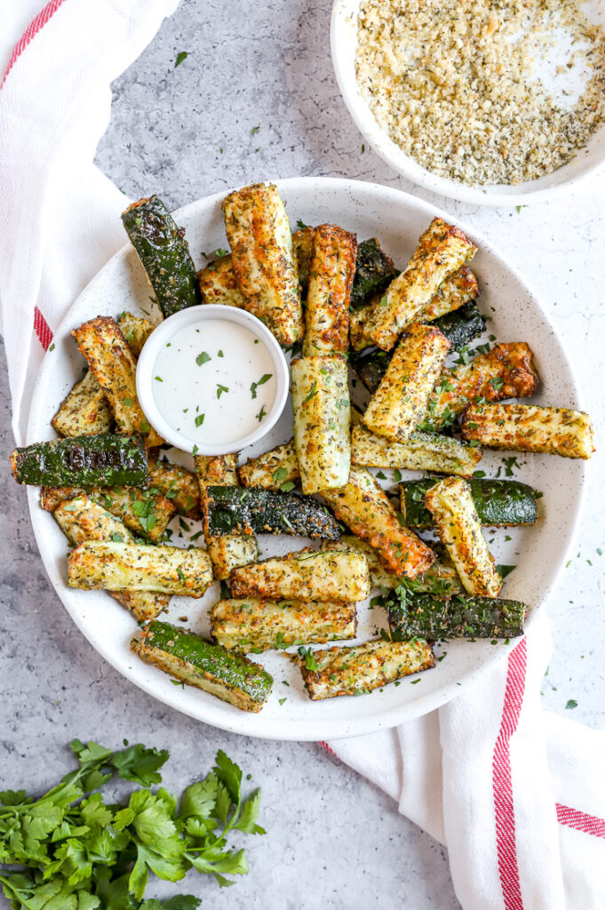 air fried zucchini fries in a large white bowl with a small bowl of ranch next to a fresh bunch of parsley and bowl of topping mixture