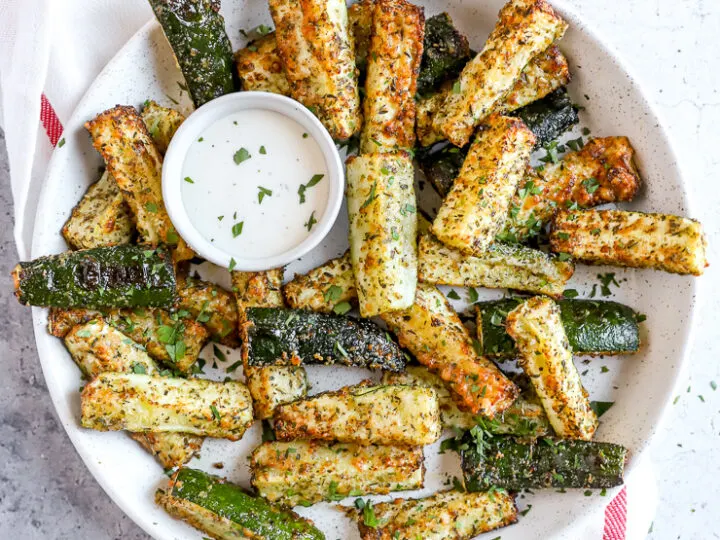 air fried zucchini fries in a large white bowl with a small bowl of ranch next to a fresh bunch of parsley and bowl of topping mixture