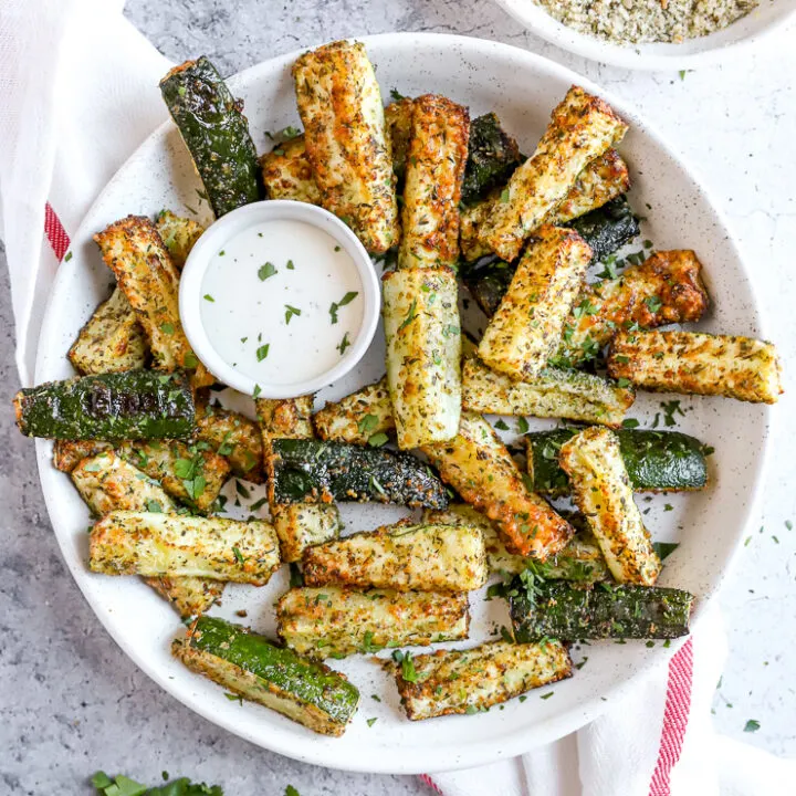 air fried zucchini fries in a large white bowl with a small bowl of ranch next to a fresh bunch of parsley and bowl of topping mixture