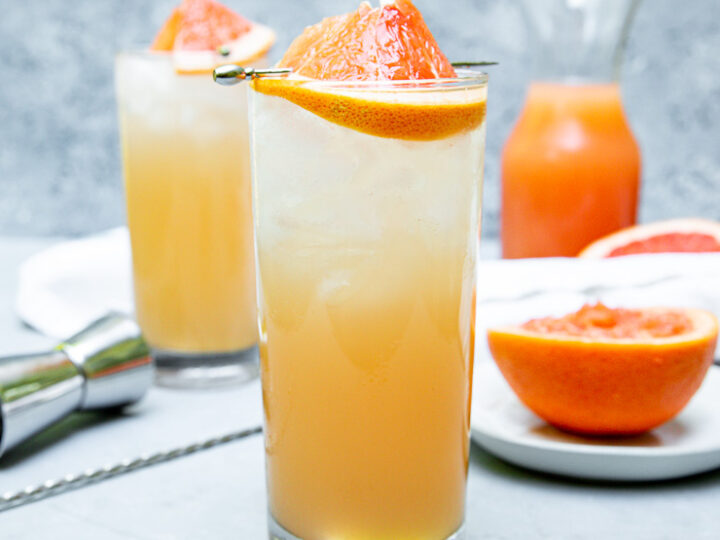 a highball glass filled with light orange grapefruit crush and a grapefruit wedge on a cocktail pick