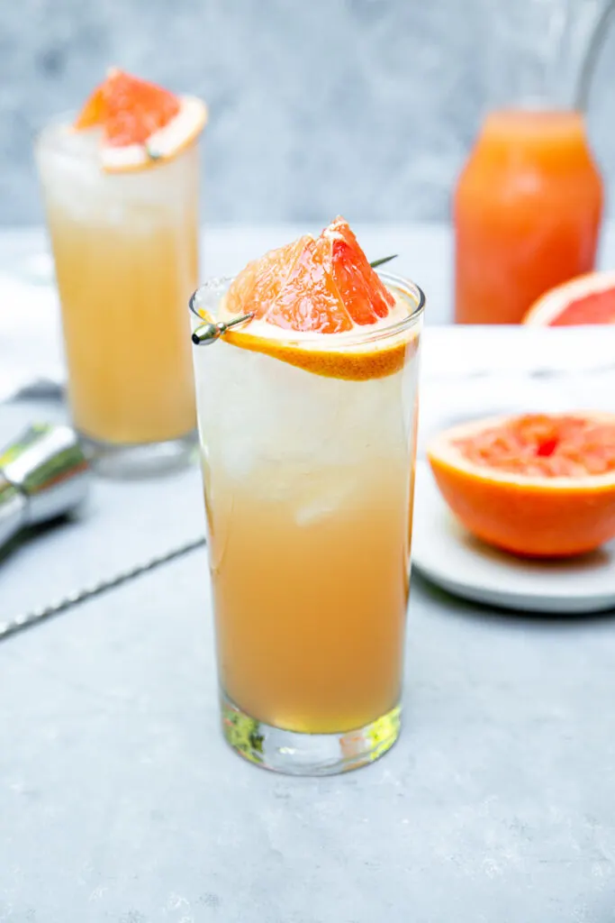 two highball glasses filled with ice and light orange grapefruit crushes surrounded by an grapefruit half and pitcher of juice