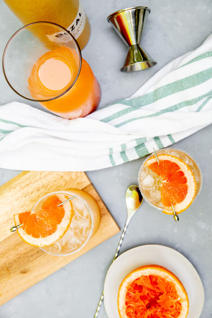 an overhead shot of two palomas, a grapefruit half, grapefruit juice, and a mezcal bottle