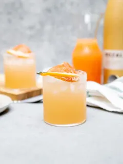 a mezcal paloma with a grapefruit slice on a cocktail pick in front of grapefruit juice in a glass pitcher