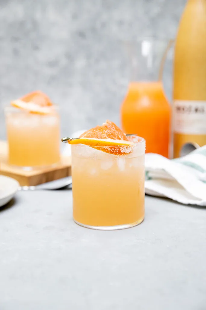 a mezcal paloma with a grapefruit slice on a cocktail pick in front of grapefruit juice in a glass pitcher