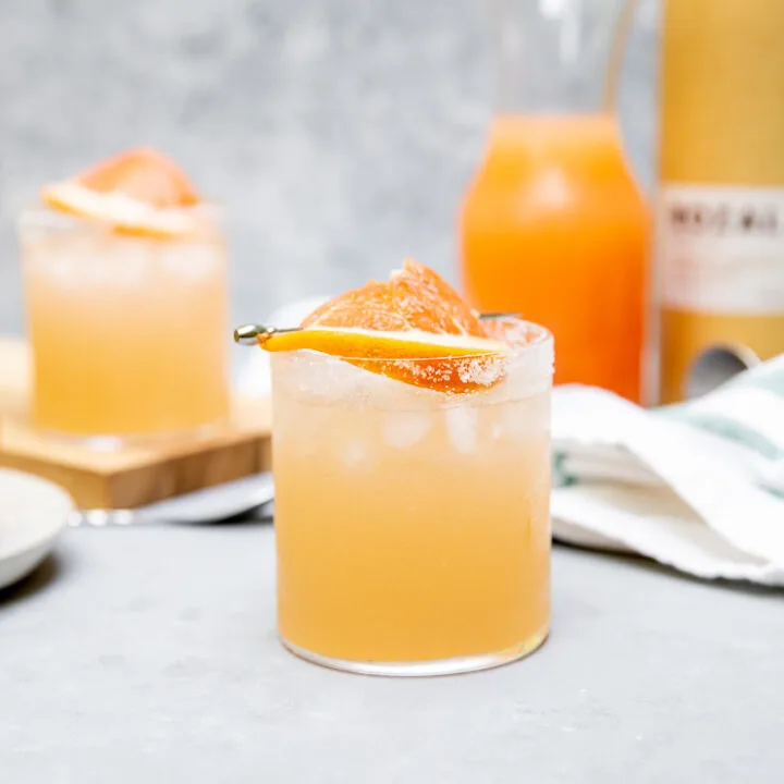 a mezcal paloma with a grapefruit slice on a cocktail pick in front of grapefruit juice in a glass pitcher