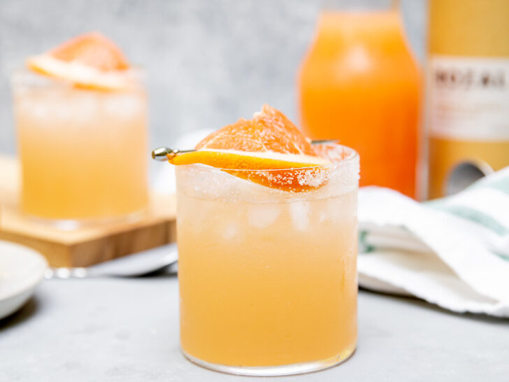 a mezcal paloma with a grapefruit slice on a cocktail pick in front of grapefruit juice in a glass pitcher