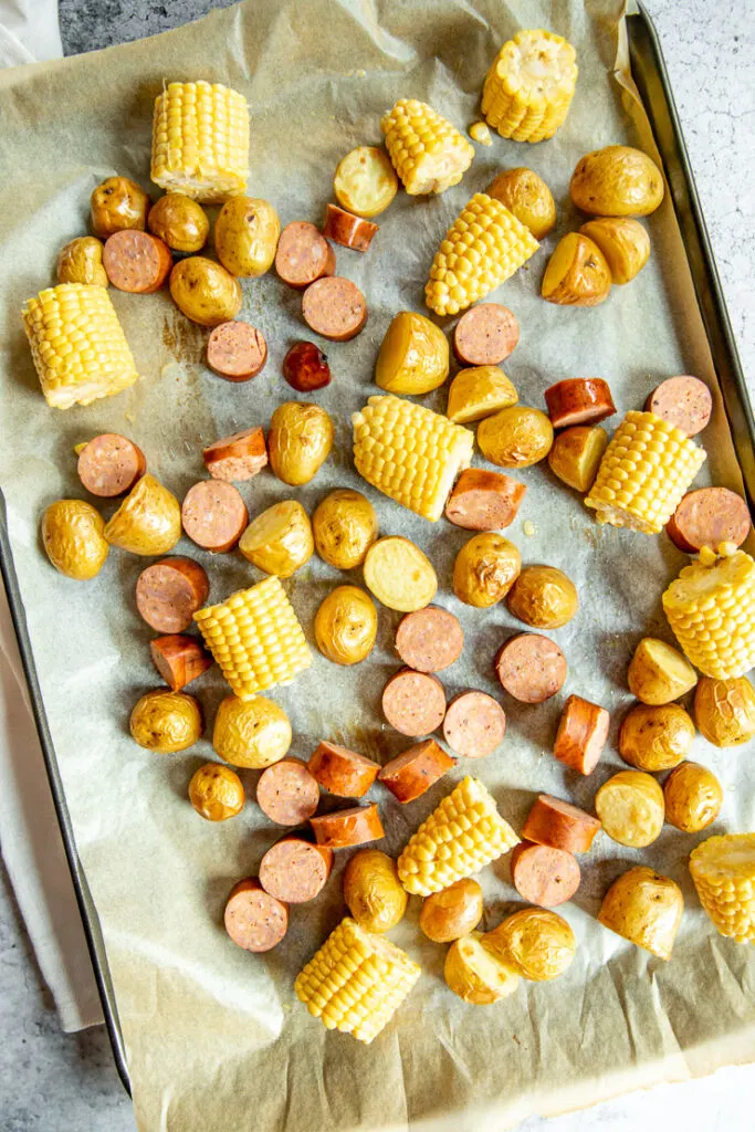 parchment paper on a cookie sheet with potatoes, sliced andouille sausage, and sliced corn