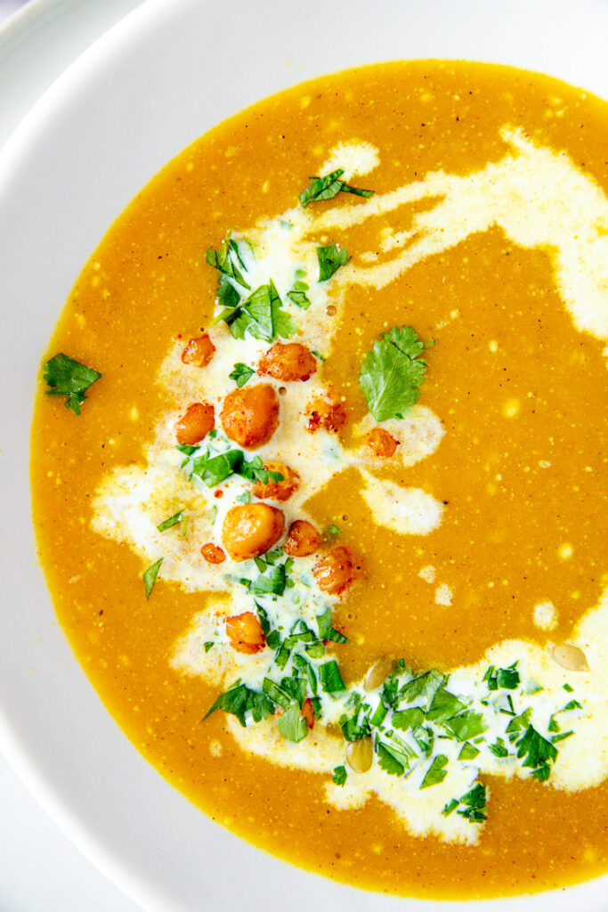 a white bowl on a white plate filled with butternut squash soup topped with a drizzle of coconut milk, diced cilantro, roasted chickpeas, and pepitas