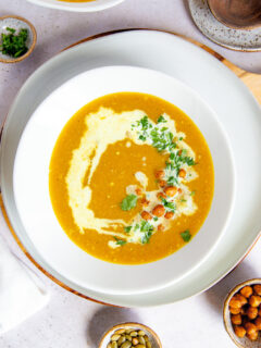 a white bowl on a white plate filled with butternut squash soup topped with a drizzle of coconut milk, diced cilantro, roasted chickpeas, and pepitas surrounded by a few small bowls filled with the toppings